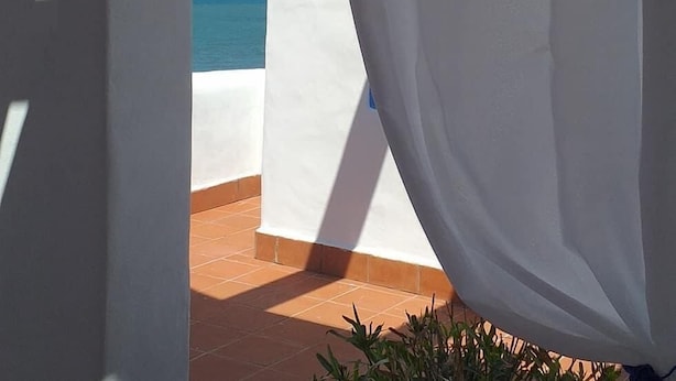 Vista a través de una cortina blanca que da a una terraza con suelo de baldosas y un cielo azul despejado.