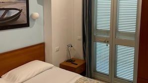 Habitación de hotel con cama, mesita de noche, lámpara, ventana con persianas y un cuadro en la pared.