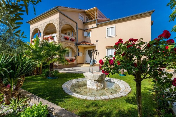 A two-story building with a stone facade, a central courtyard with a fountain, and a well-maintained garden with palm trees and flowering plants.