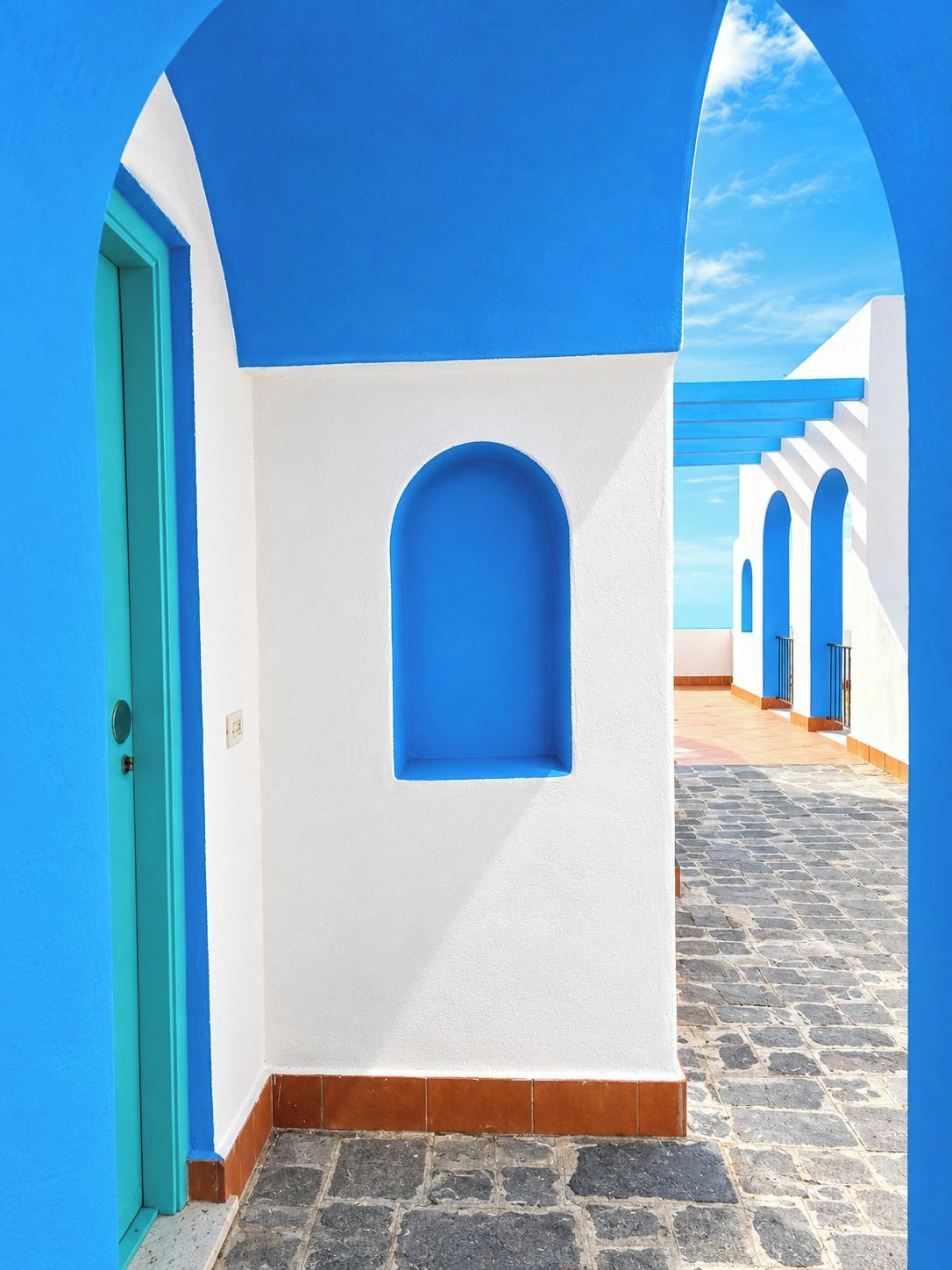 Un stretto vicolo con una porta blu, un muro bianco con una nicchia blu e un sentiero lastricato in pietra che conduce a un edificio bianco con dettagli blu.