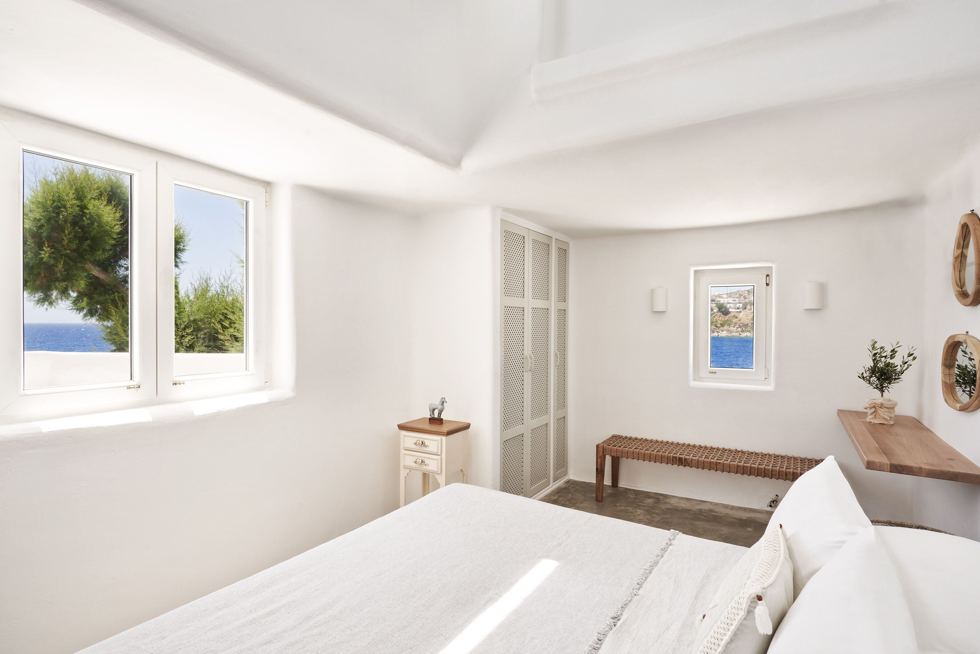 A bedroom with a bed, a wooden bench, a small table, a wardrobe, and a window with a view of the sea.