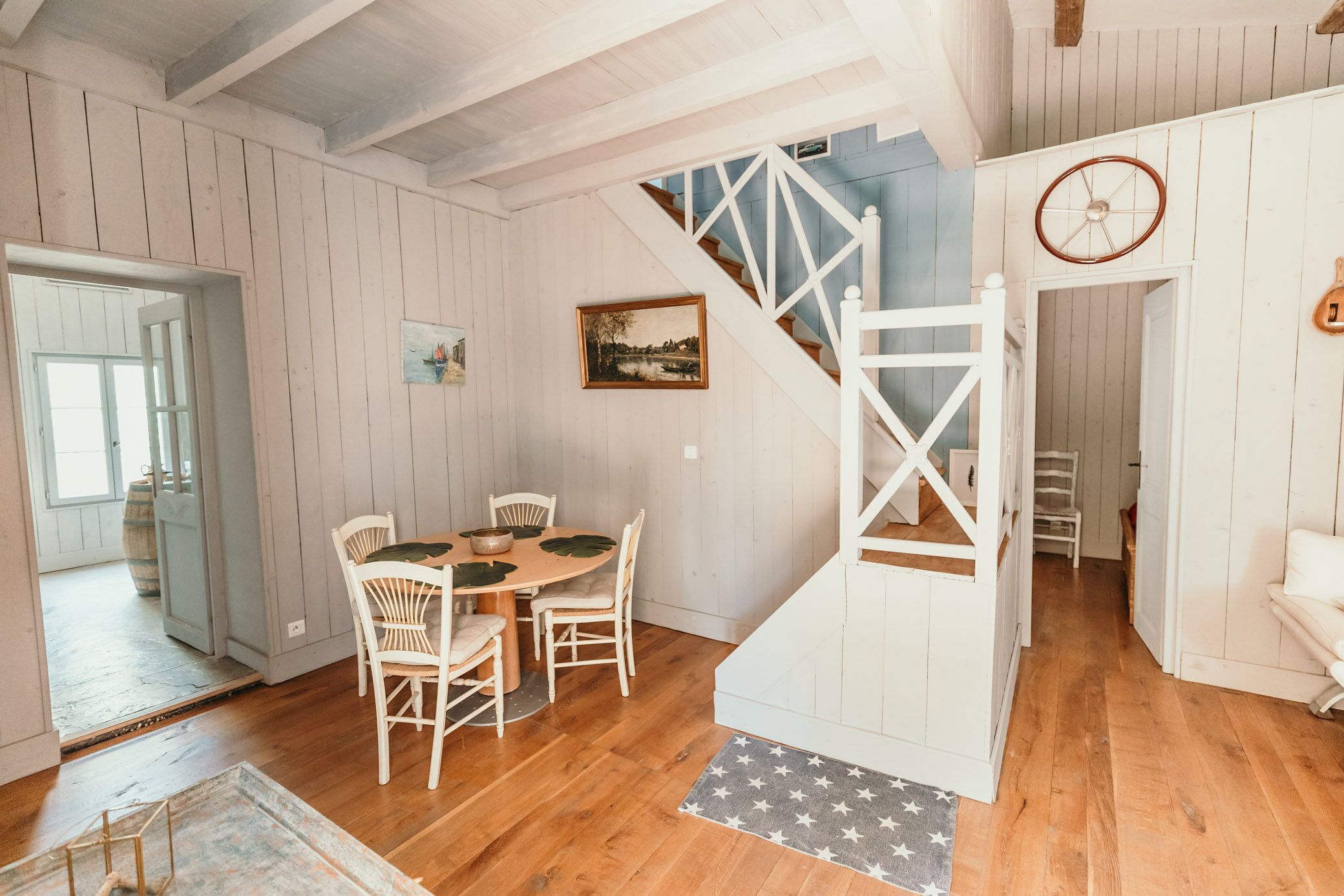 Un coin repas avec une table et des chaises en bois, un escalier avec une rampe blanche et un tableau accroché au mur.