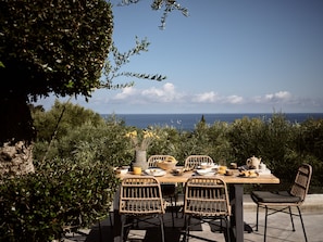 An outdoor dining table with wicker chairs, set against a backdrop of a clear sky and ocean view.