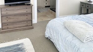 Un dormitorio con cama, televisor sobre un aparador de madera, baño con ducha y mesita de noche con lámpara.