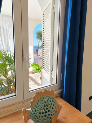 A room with a view of a courtyard, a wooden table with a decorative fish, and a door with white shutters.