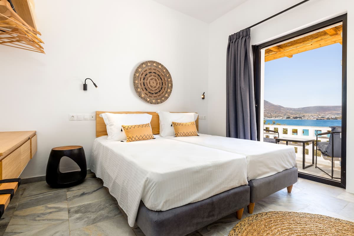 Ein Schlafzimmer mit einem Bett, einem kleinen Hocker, einer Wandleuchte und Meerblick vom Balkon.