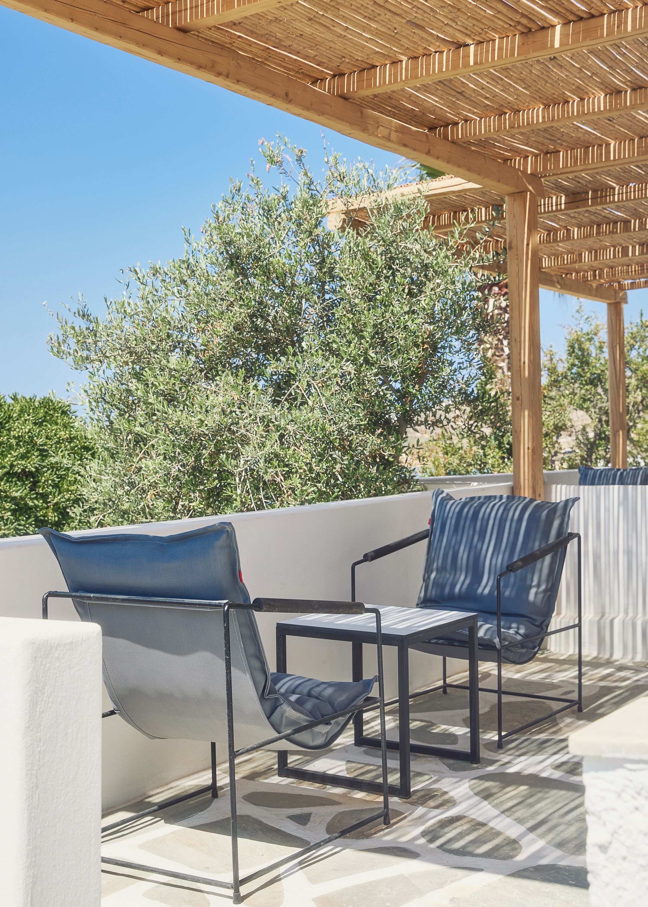 An outdoor seating area with two chairs and a table, a pergola overhead, and a view of trees and a building in the background.