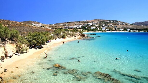 Uma praia costeira com águas turquesa cristalinas, litoral rochoso e edifícios brancos ao fundo.
