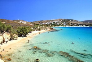 A coastal beach with clear turquoise waters, rocky shoreline, and white buildings in the background. - Omilos Beachfront House (Paros)