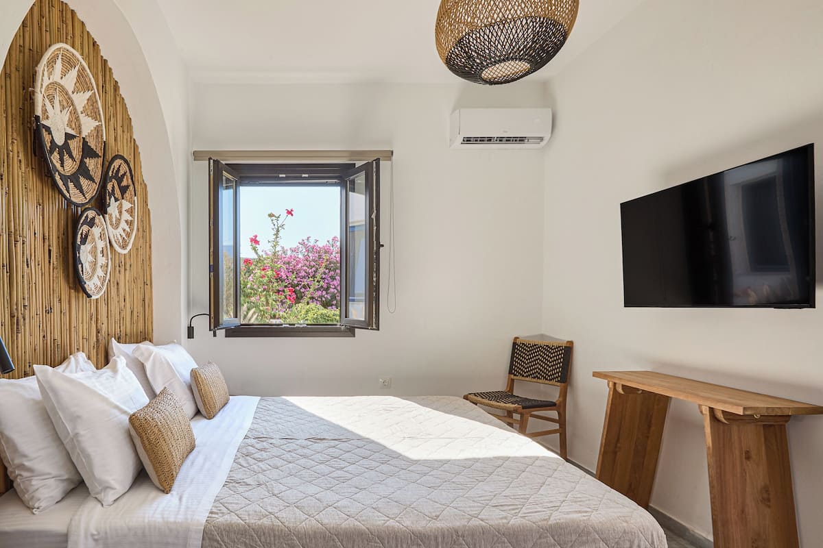 Ein Schlafzimmer mit Bett, Fernseher, Fenster mit Blick auf Blumen und einer Wandklimaanlage.
