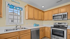 Uma cozinha com armários de madeira, eletrodomésticos de aço inoxidável, lava-louças e uma janela com cortina decorada com tema praiano.