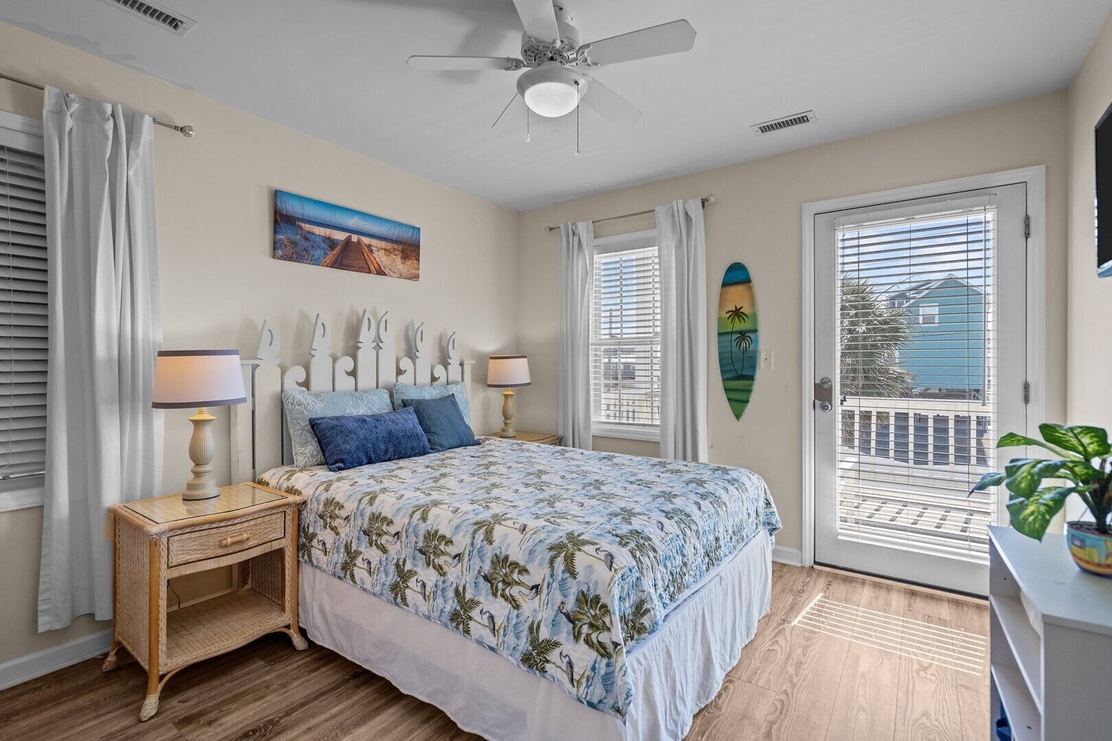 A bedroom with a bed, two bedside tables, a ceiling fan, a window with blinds, a door, and a view of the outside.