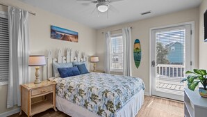 Quarto com cama, duas mesas de cabeceira, ventilador de teto, janela com persianas, porta e vista para o exterior.