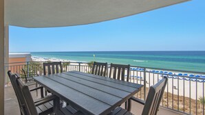 A balcony with a table and chairs, overlooking a beach with umbrellas and a flag. - 302 -Luxury Gulf-Front Condo | 3 Bedrooms | Sleeps 8+ Free Beach Chairs! (Panama City Beach)