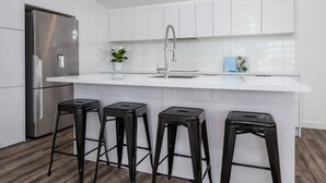 Eine moderne Küche mit weißen Schränken, einem Edelstahlkühlschrank, einer Spüle mit einer hohen Armatur und drei schwarzen Barhocker.