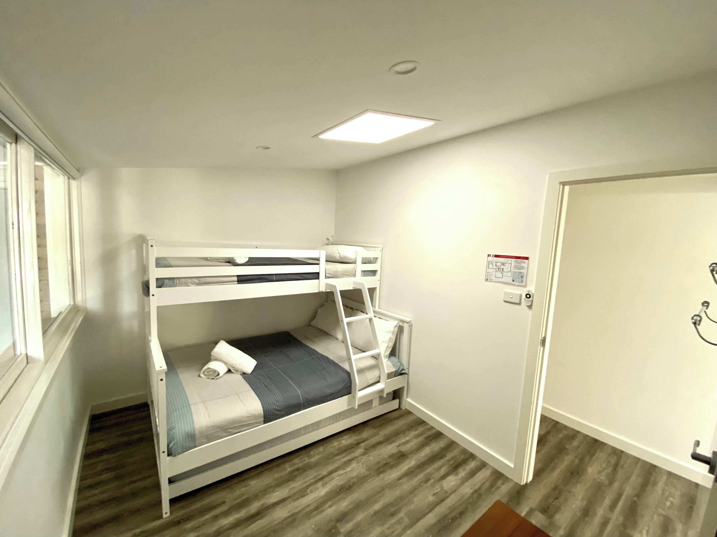 Une chambre avec un lit superposé, une fenêtre, une porte et un interrupteur sur le mur.