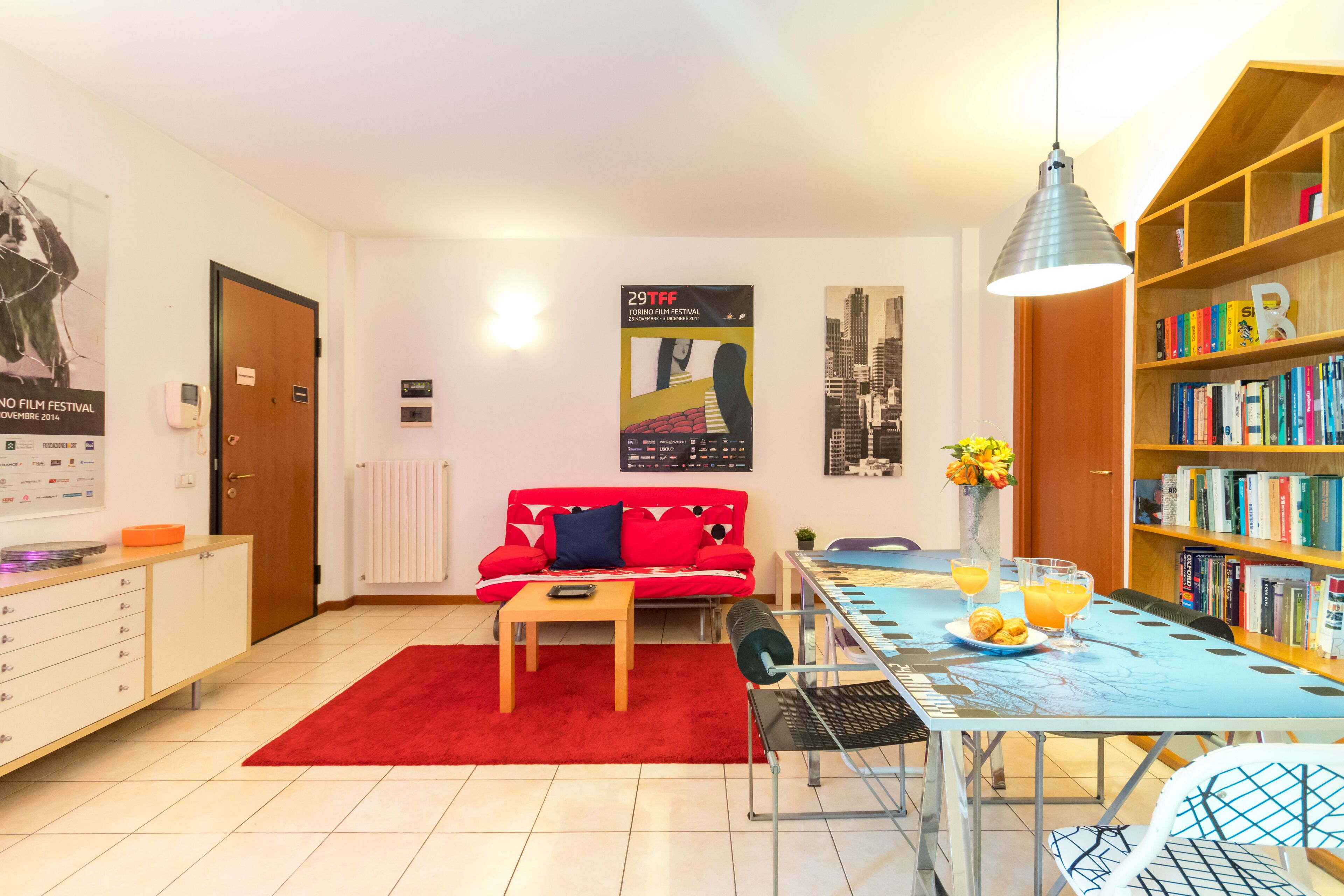 Uma sala de estar moderna com um sofá vermelho, uma mesa de jantar com toalha azul e uma estante repleta de livros.