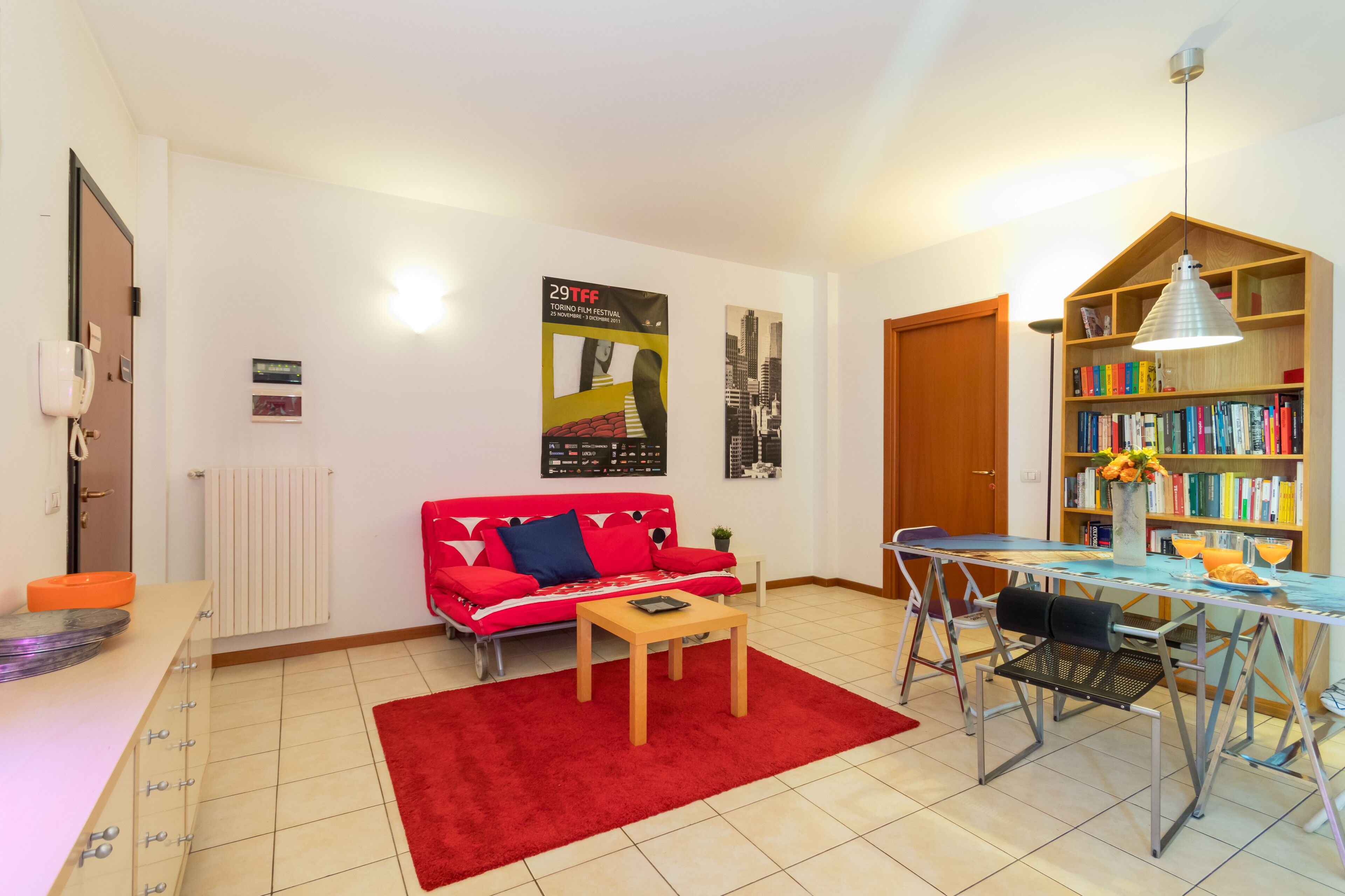 Una sala de estar con un sofá rojo, una mesa de centro, una mesa de comedor con sillas, una estantería y un radiador.
