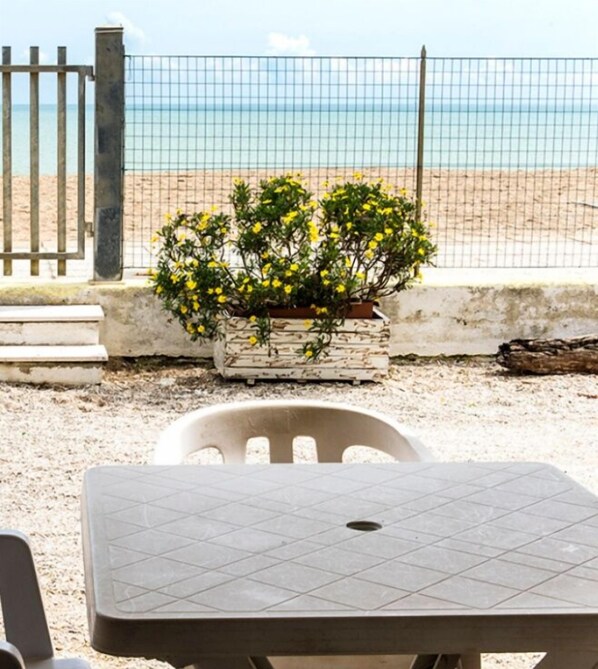 A table with a grid pattern, a potted plant with yellow flowers, and a view of a beach through a fence.