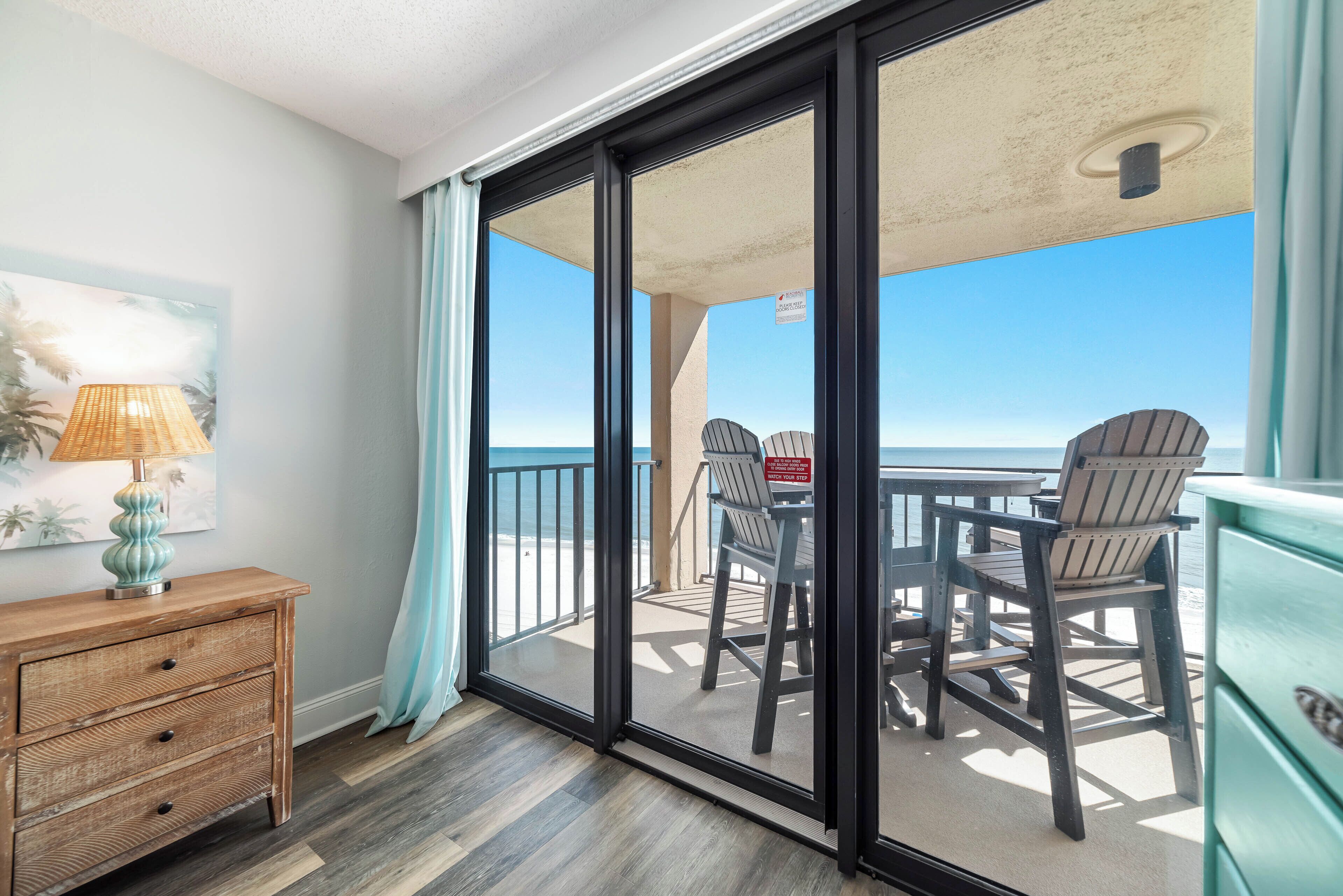 A balcony with a view of the beach, a wooden table and chairs, and a lamp on a dresser.