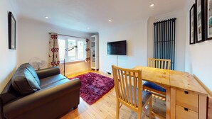 Una sala moderna con sofá, mesa de comedor y sillas. Hay un televisor montado en la pared y una ventana con cortinas.