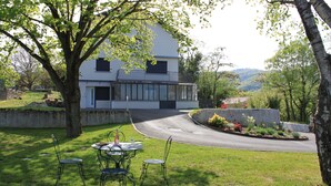 Ein modernes Haus mit einer geschwungenen Auffahrt, einem Essbereich mit Gartenmöbeln und einem großen Baum im Vordergrund.