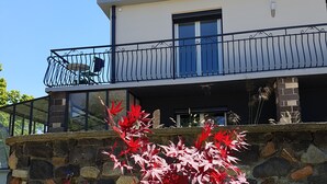 Ein zweistöckiges Haus mit einer Stein-Fassade, einem Balkon mit Metallgeländer und einem kleinen Garten mit roten Blättern.