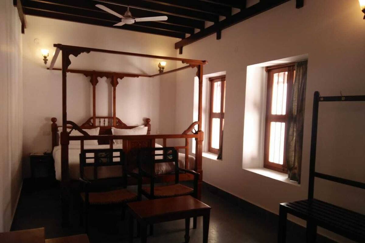 A room with a four-poster bed, wooden chairs, a table, and a window with curtains.