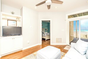 A modern living room with a flat-screen TV, a ceiling fan, and a large window offering a view of the beach.