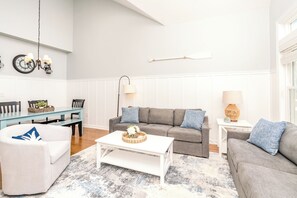 A living room with a grey sofa, white armchair, and a coffee table. There is a dining table with chairs and a wall clock. - Feeling TipSea - 3 BR Townhome - Amazing Ocean Views & Community Pool (Surf City)