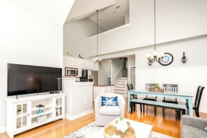 A modern living space with a flat-screen TV, kitchen appliances, a dining area with a bench, and a staircase leading to an upper level.