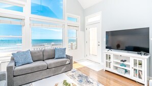 Een woonkamer met een grote televisie, een grijze bank en uitzicht op het strand.