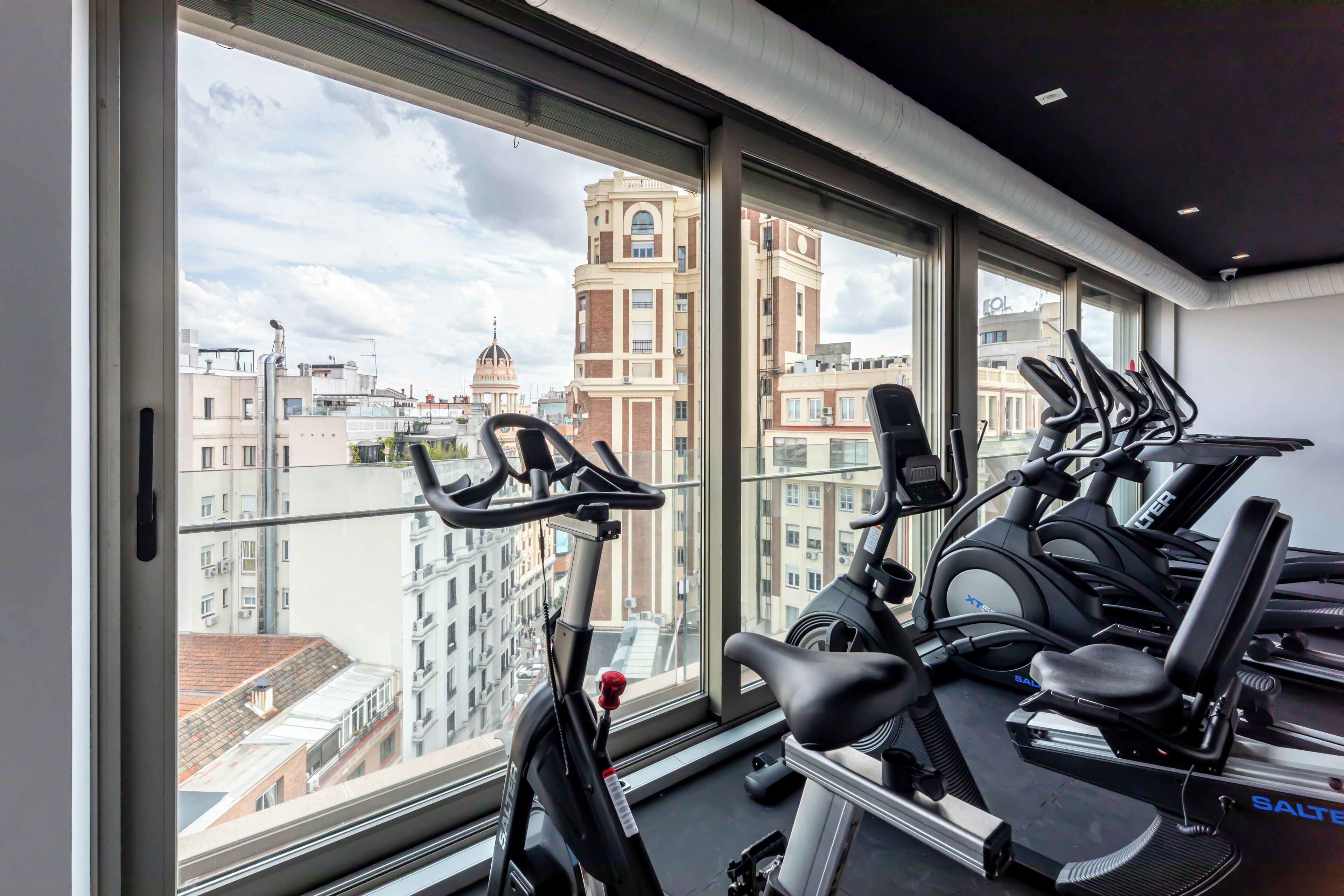 Ein Fitnessraum mit Ergometer und Laufband, großen Fenstern mit Blick auf die Stadt sowie einem modernen Deckenstil.