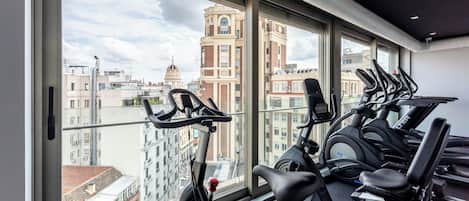 Academia com bicicletas ergométricas e esteira, amplas janelas com vista para a cidade e design moderno no teto.