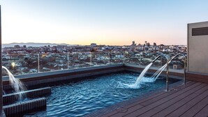 A rooftop pool with two water jets, surrounded by a cityscape view at dusk. - Gran View Apartments (Madrid)