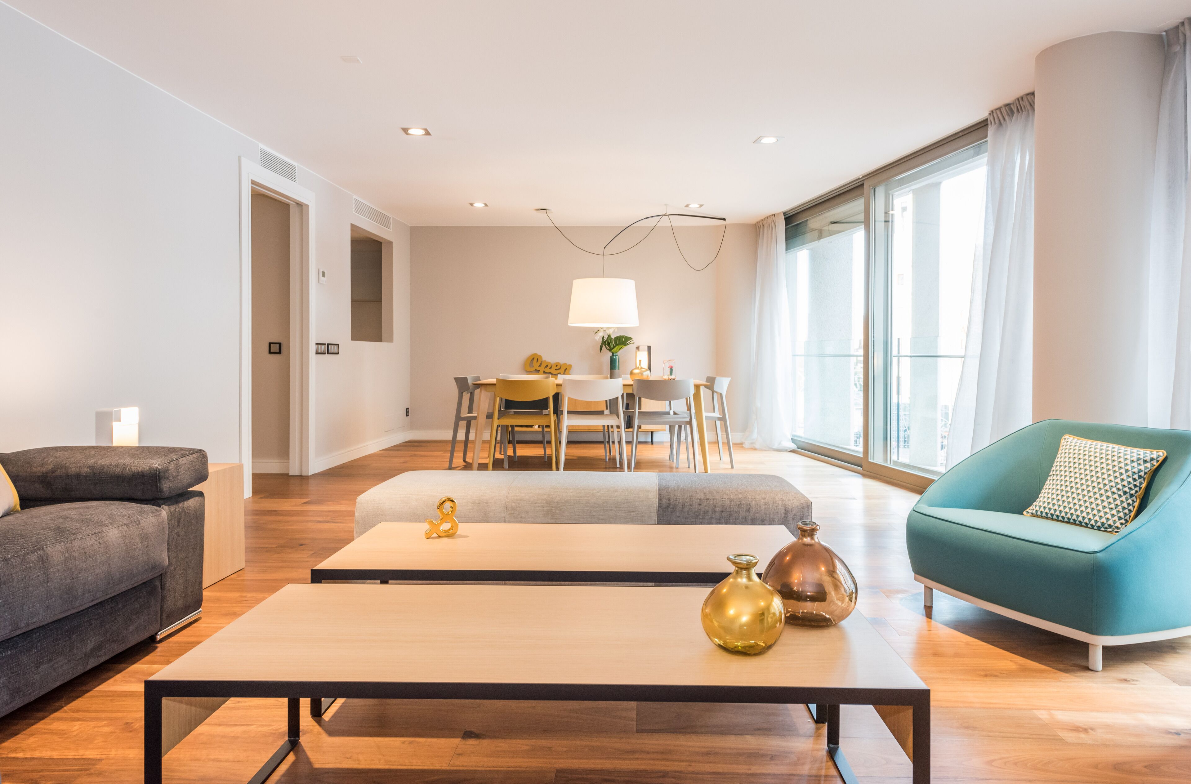 A modern living room with a sofa, a coffee table, and a teal armchair. There is a dining area with a table and chairs in the background.