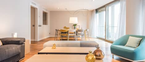 A modern living room with a sofa, a coffee table, and a teal armchair. There is a dining area with a table and chairs in the background.