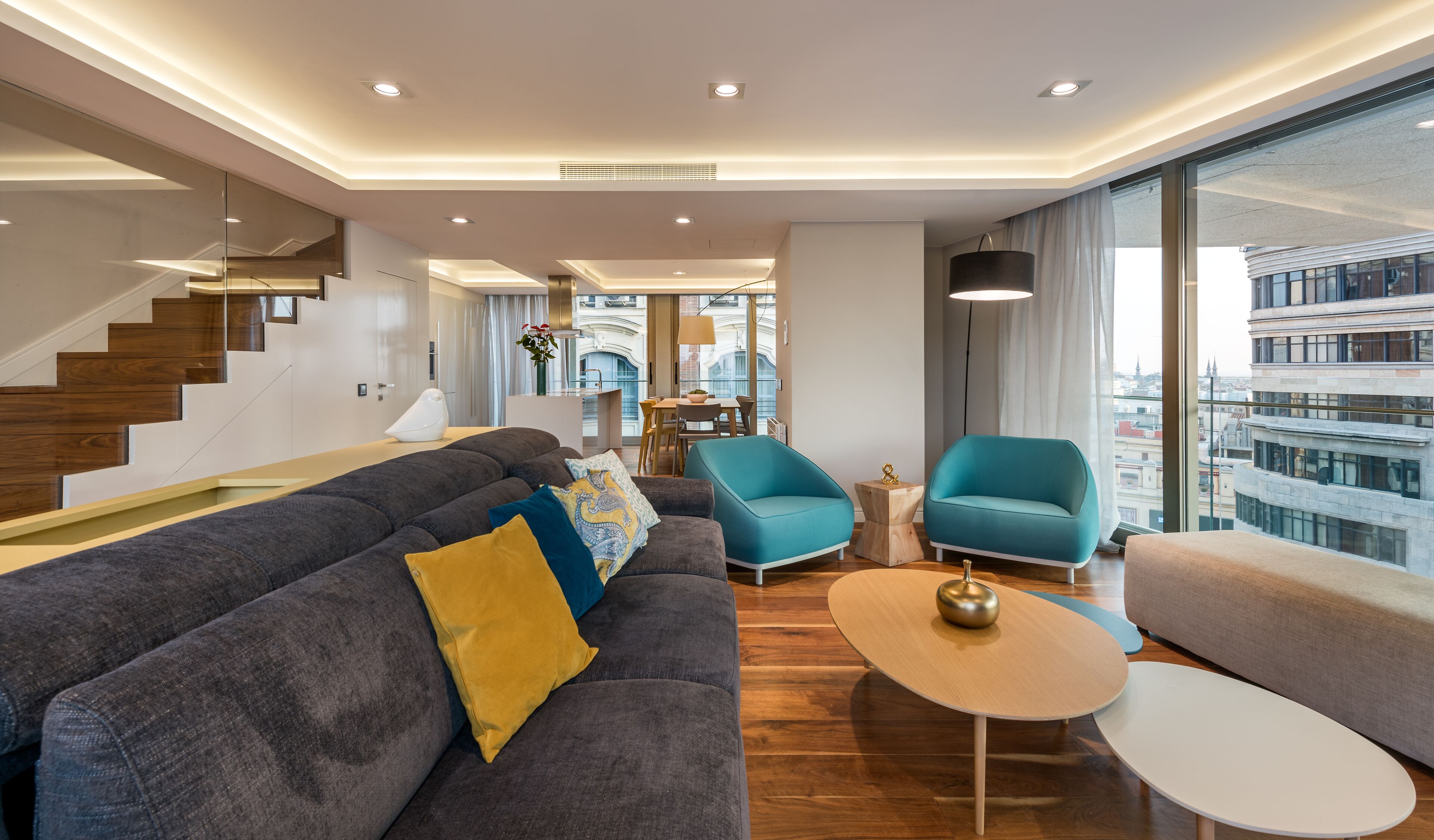 A modern living room with a grey sofa, a teal armchair, and a wooden coffee table. A staircase with wooden steps leads to an upper level.