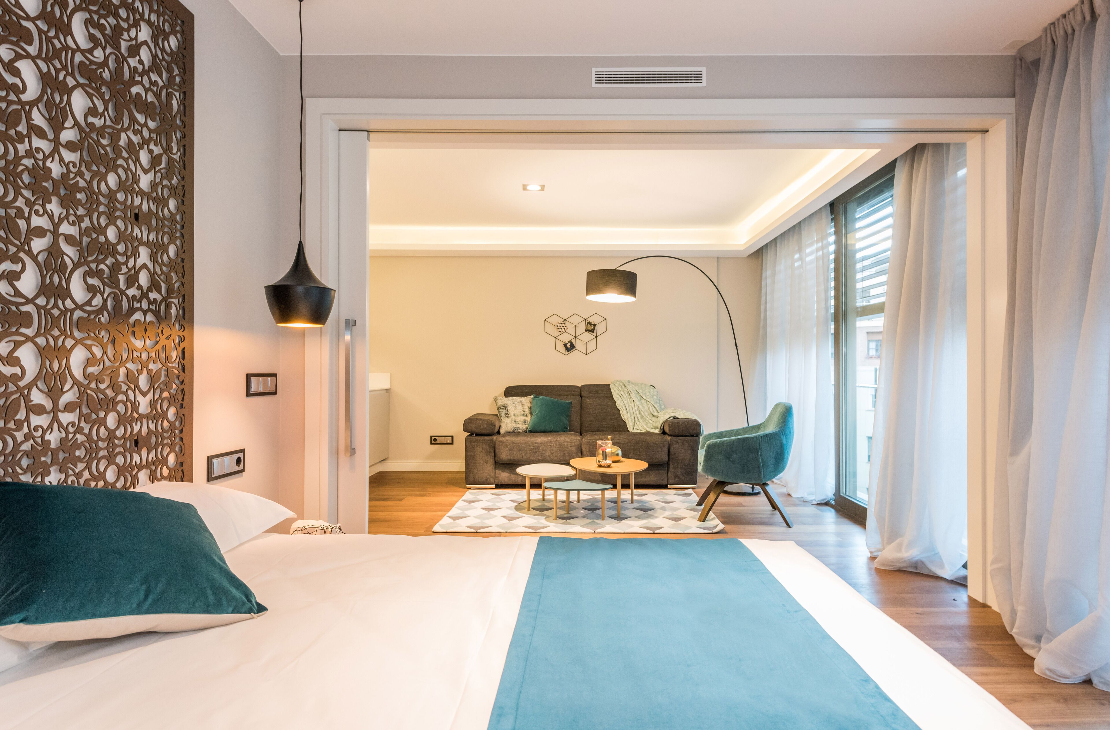 Une chambre d’hôtel moderne avec un grand lit, un canapé, une table basse et un fauteuil. On y trouve un panneau décoratif au mur et une fenêtre avec des rideaux.