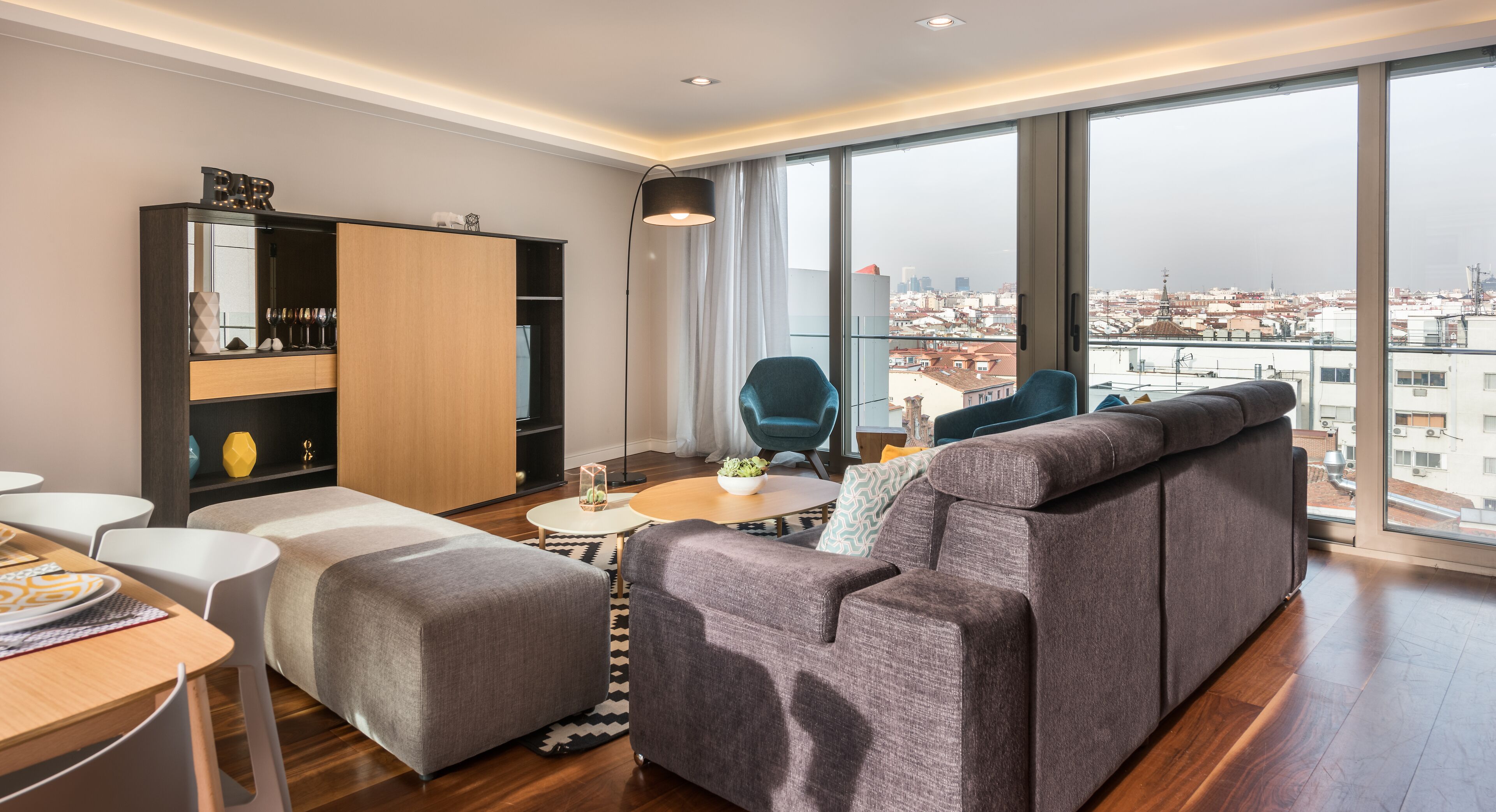 Un salon moderne avec un canapé, un pouf et un coin repas comprenant une table et des chaises. Une grande fenêtre offre une vue sur la ville.