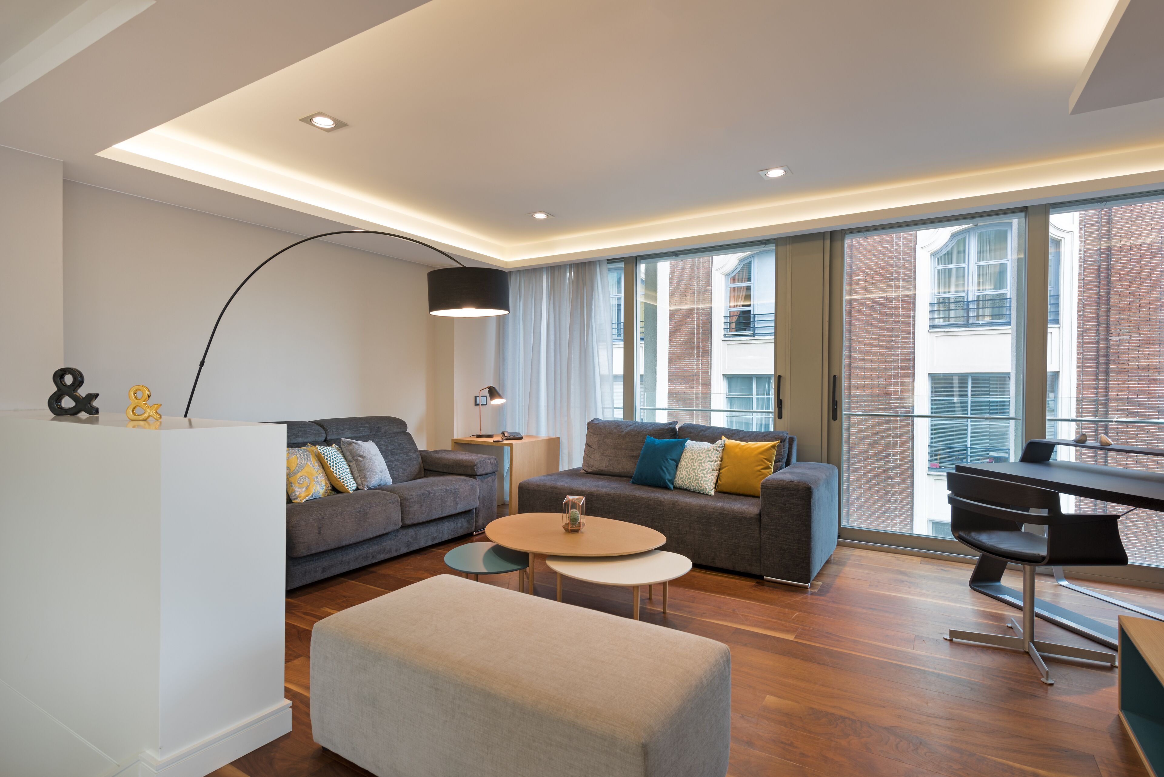 Una sala de estar moderna con sofá, reposapiés y mesa de centro, un escritorio y ventanas amplias con vistas a edificios.