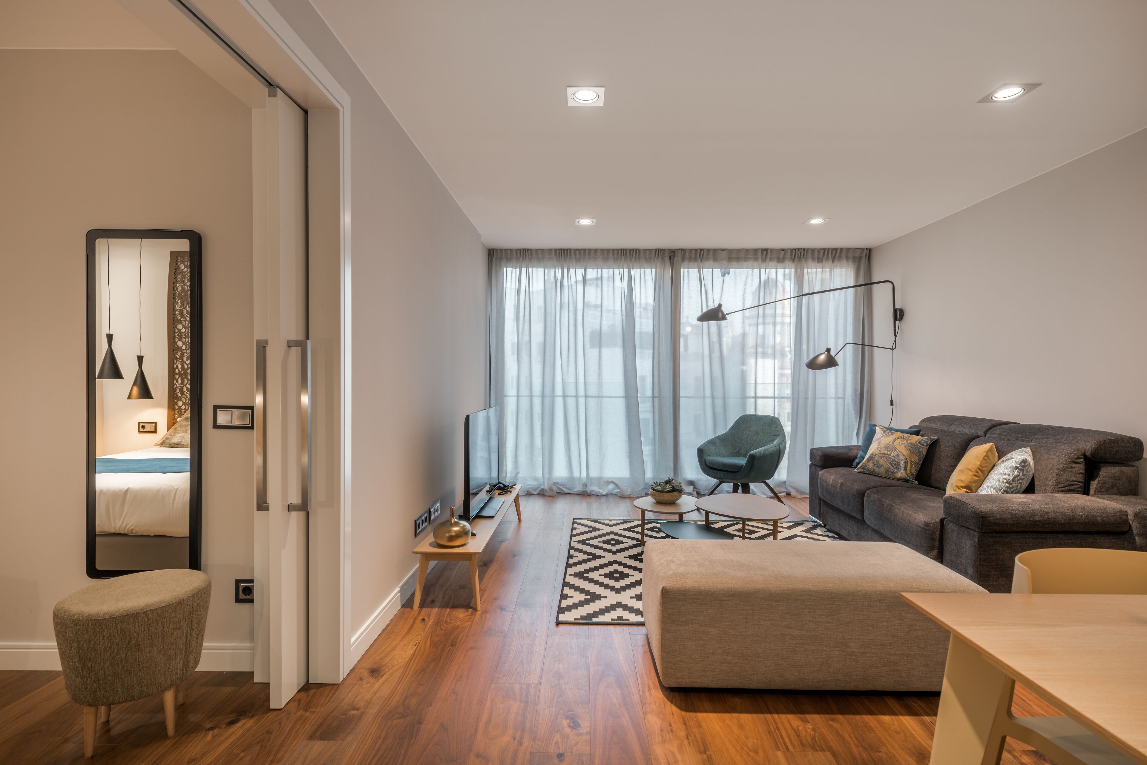 Een moderne woonkamer met een bank, voetenbankje en een eettafel. Er is een televisie, een lamp en een plant op een bijzettafeltje.