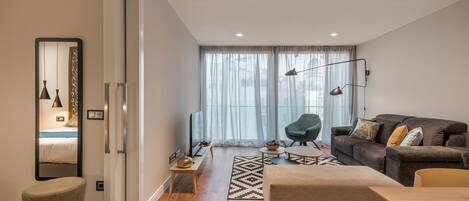 A modern living room with a sofa, ottoman, and a dining table. There is a TV, a lamp, and a plant on a side table.
