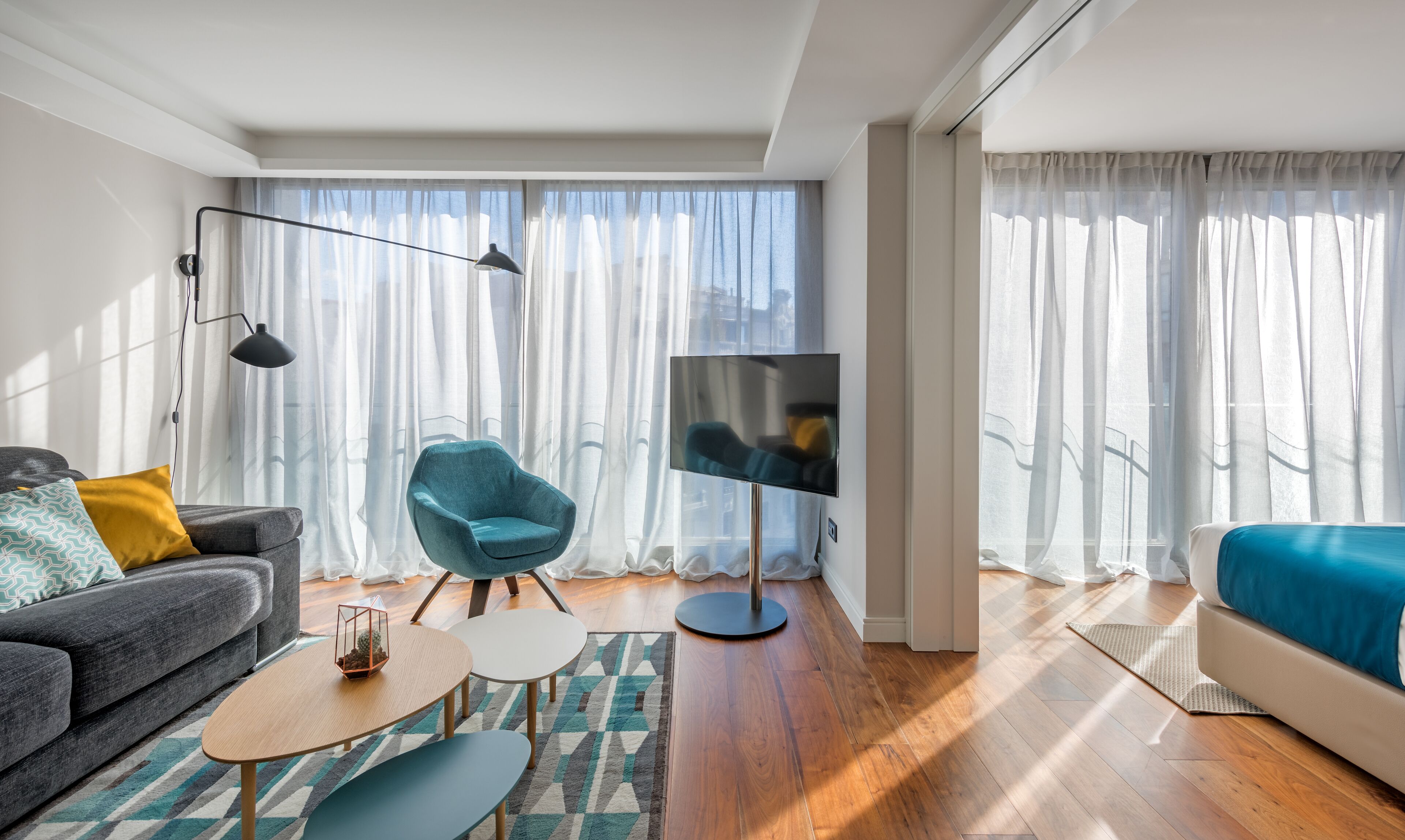 Una sala de estar moderna con un sofá, un sillón azul, un televisor y ventanas grandes con cortinas translúcidas.