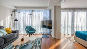 Una sala de estar moderna con un sofá, un sillón azul, un televisor y ventanas grandes con cortinas translúcidas.
