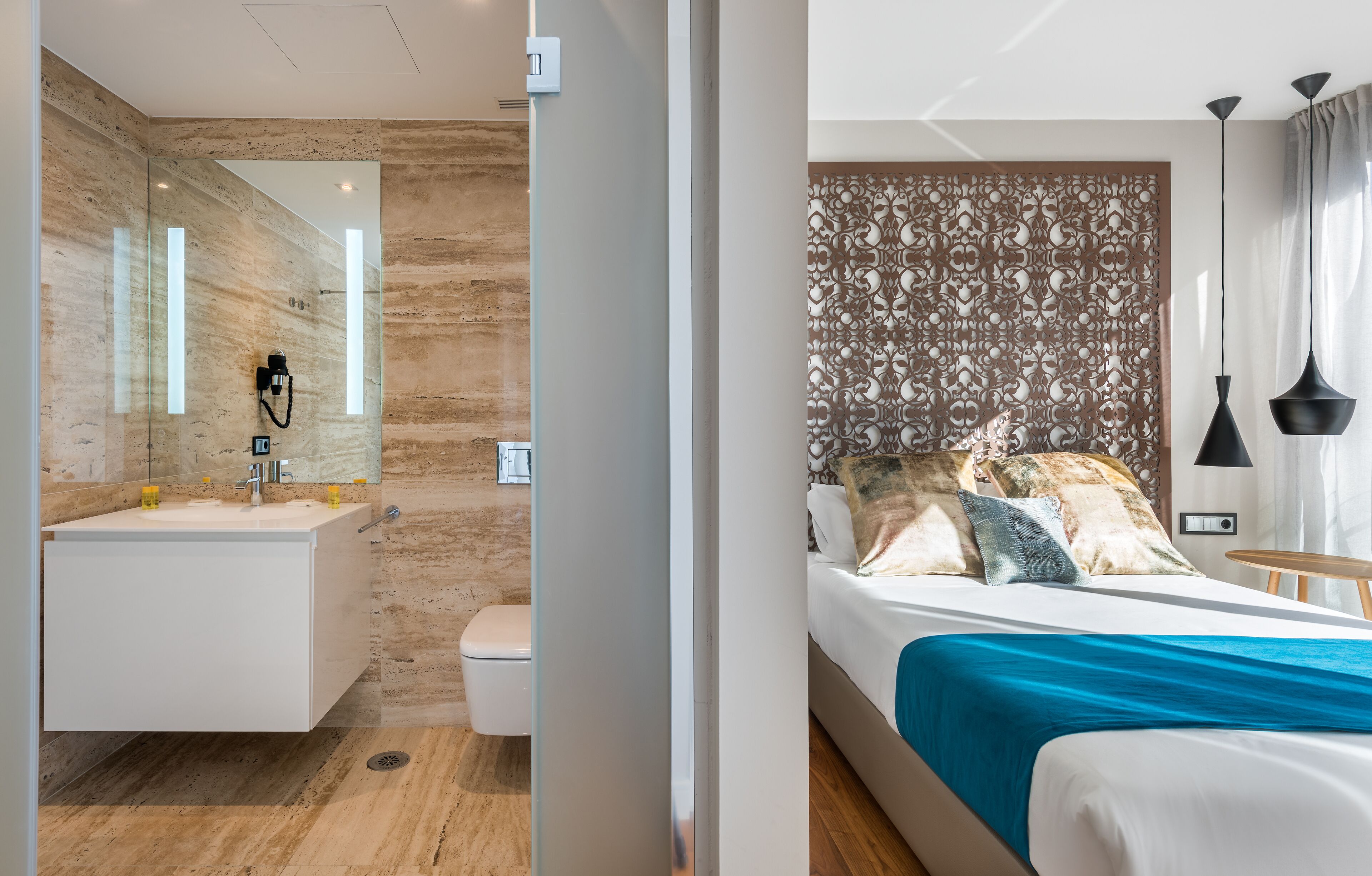 A modern bathroom with a marble sink, toilet, and shower. The bedroom features a bed with a blue bedspread, decorative pillows, and a patterned headboard.