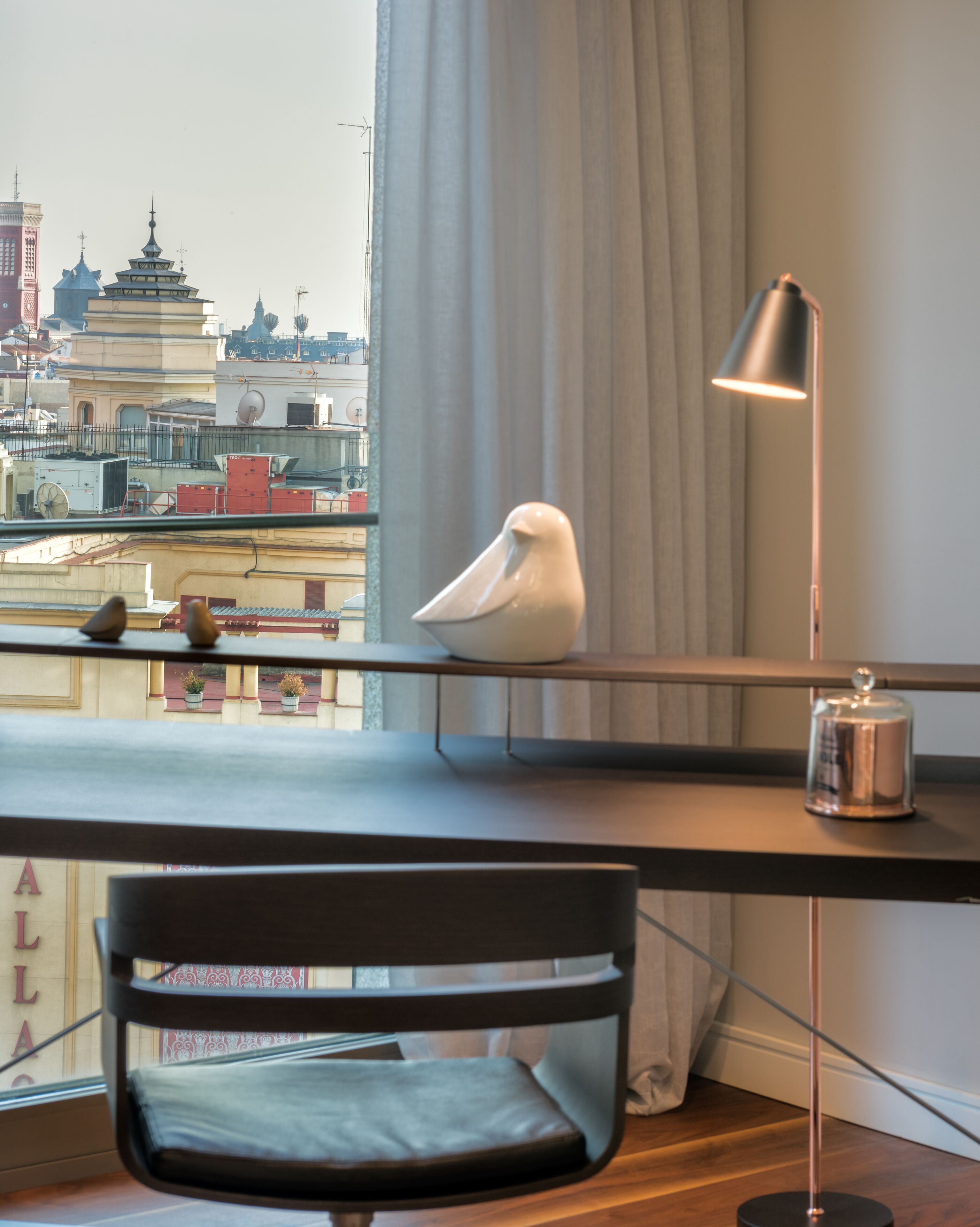 A modern room with a desk, a chair, a decorative bird, a lamp, and a view of the cityscape through a window.