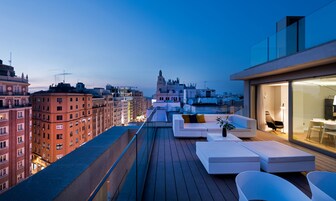 A rooftop terrace with modern outdoor seating, a view of the cityscape, and a building with a tower in the background.