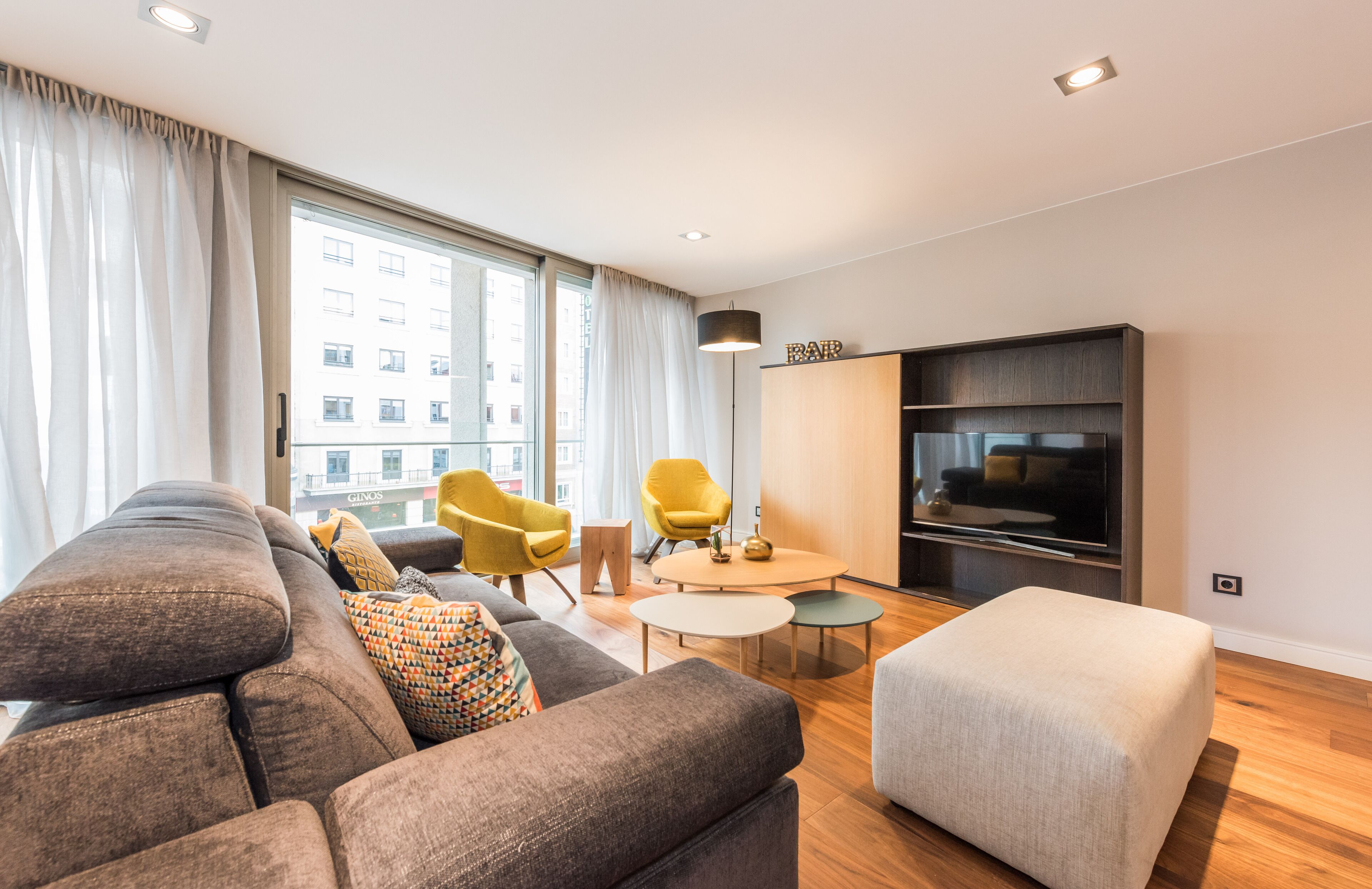 A modern living room with a large sofa, two yellow chairs, a small round table, and a flat-screen TV mounted on the wall.