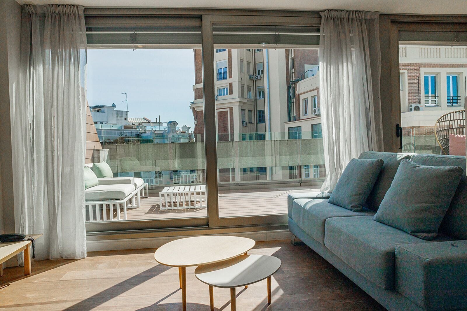 A balcony with a sofa and two tables, overlooking a cityscape with buildings and boats.